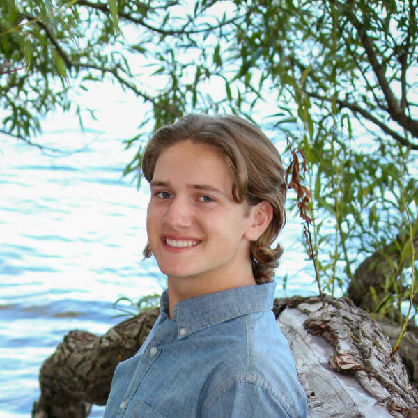 Daniel Smith, an undergraduate research assistant in the Sleep and Health Research Lab. He is smiling, wearing a blue chambray shirt, while leaning against a tree branch near a body of water.