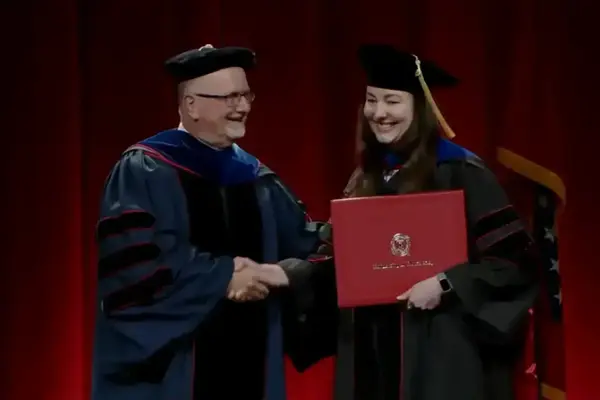 Smiling man in dark blue faculty regalia shakes hands with a smiling woman in black cap and gown holding a red diploma.